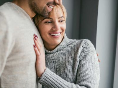 Angebote- Paarberatung-Ein junges Paar steht am Fenster. Die Frau hat ihren Kopf auf der Schulter des Partners. Beide lächeln zuversichtlich.