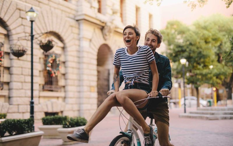 Ein Freundlicher Sommertag. Ein junges Parr sitz auf dem Fahrrad und fährt lachend und glücklich durch die Stadt. 