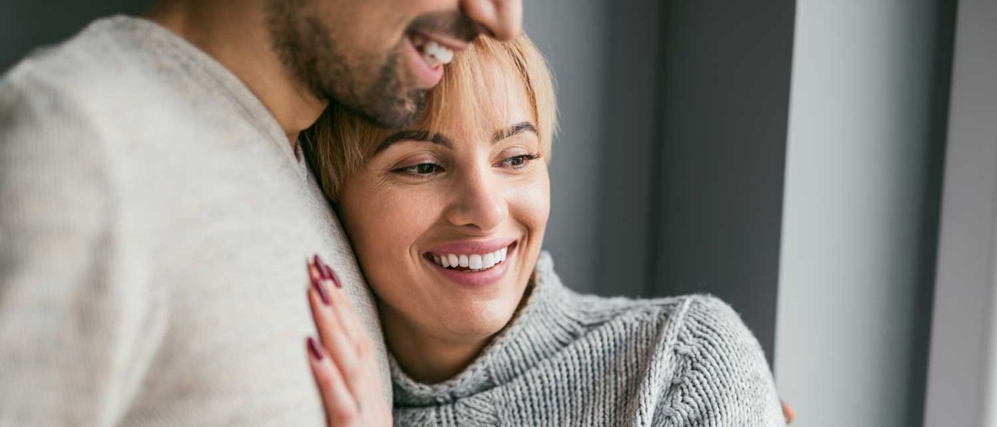 Paarberatung. Ein junges Paar steht am Fenster. Die Frau hat ihren Kopf auf der Schulter des Partners. Beide lächeln zuversichtlich. Paarberatung kann dabei helfen.